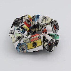 May include: A white scrunchie with a colourful pattern of vintage cameras. The scrunchie is made of soft fabric and is perfect for holding hair in place.