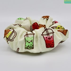 以下が含まれることがあります： カラフルな漫画のタピオカミルクティーのカップの模様がついた白い布製のシュシュ。カップは笑顔で、さまざまな色のタピオカで満たされています。