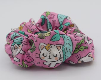 Könnte beinhalten: Ein rosa Stoff-Haargummi mit einem Meerjungfrauen-Katzen-Druck. Der Druck zeigt eine weiße Katze mit geschlossenen Augen, einer gelben Blume und einem grünen Meerjungfrauen-Schwanz.