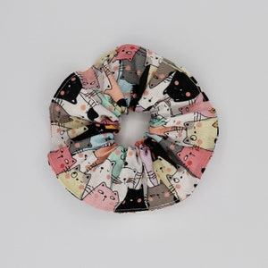 May include: A colorful scrunchie featuring a repeating pattern of cartoon cats in various colors, including black, pink, and gray, on a white background. The fabric is gathered around an elastic band, creating a ruffled effect. A fun hair accessory.