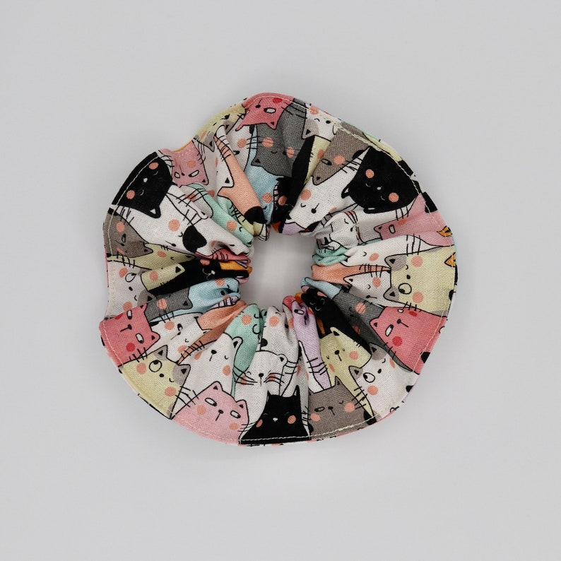 May include: A colorful scrunchie featuring a repeating pattern of cartoon cats in various colors, including black, pink, and gray, on a white background. The fabric is gathered around an elastic band, creating a ruffled effect. A fun hair accessory.