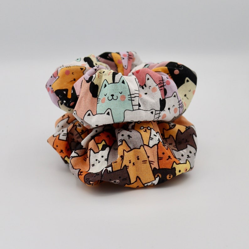 May include: Two colorful scrunchies stacked on top of each other. The fabric is patterned with a repeating design of cartoon cats in various colors, including orange, brown, white, and pastel shades. The scrunchies are made of soft fabric.