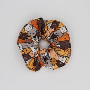 May include: A fabric scrunchie featuring a repeating pattern of cartoon cats in various colors, including brown, orange, gray, and white. The scrunchie is gathered in the center, creating a ruffled effect. This hair accessory is suitable for all hair types.