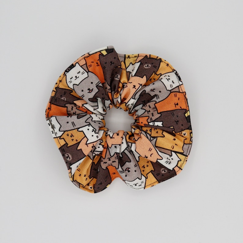 May include: A fabric scrunchie featuring a repeating pattern of cartoon cats in various colors, including brown, orange, gray, and white. The scrunchie is gathered in the center, creating a ruffled effect. This hair accessory is suitable for all hair types.