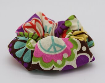 Könnte beinhalten: Ein farbenfrohes Haargummi mit einem Retro-Blumen- und Peace-Zeichen-Muster. Der Stoff weist Farbtöne von Rosa, Lila, Grün, Blau und Braun auf. Dieses Haaraccessoire eignet sich perfekt, um jeder Frisur einen Farbtupfer zu verleihen.