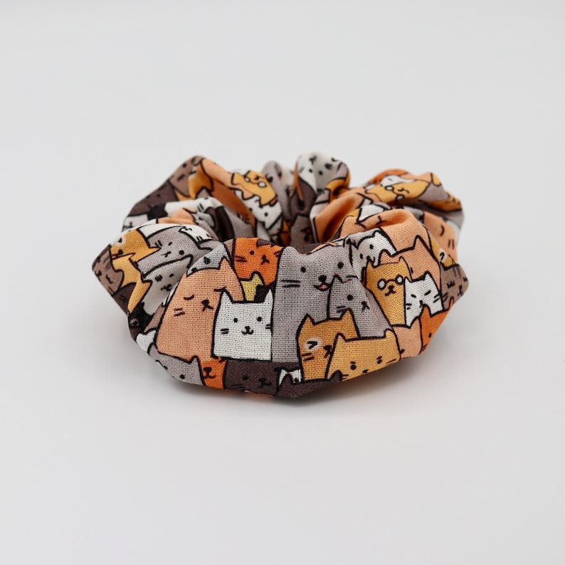 May include: A fabric scrunchie featuring a repeating pattern of cartoon cats in various colors, including orange, brown, gray, and white. The scrunchie is gathered and appears to be made of a soft material, suitable for hair styling.