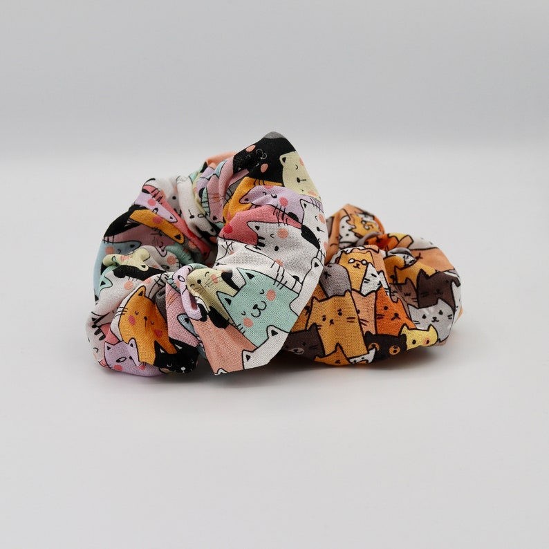 May include: Two colorful scrunchies with a repeating pattern of cartoon cats in various colors. The scrunchies are made of fabric and appear to be new. The background is plain white.