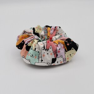 May include: A colorful scrunchie featuring a repeating pattern of cartoon cats and dogs in various colors. The fabric is white with the illustrated animals in shades of pink, yellow, black, and blue. The scrunchie is gathered and circular.