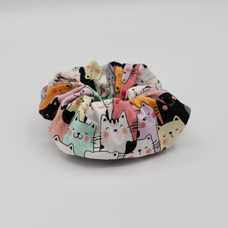 May include: A colorful scrunchie featuring a repeating pattern of cartoon cats and dogs in various colors. The fabric is white with the illustrated animals in shades of pink, yellow, black, and blue. The scrunchie is gathered and circular.