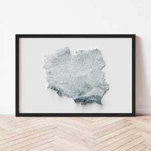 May include: A framed, three-dimensional map of Poland, rendered in shades of gray and blue. The map is set against a white background and enclosed in a black frame. The artwork is displayed on a light-colored wooden floor.