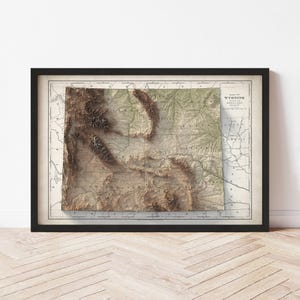 May include: Framed vintage map of Wyoming, featuring a detailed relief of the state's topography. The map is in shades of brown, beige, and green, with a black frame. The map is displayed on a white wall above a wooden floor.