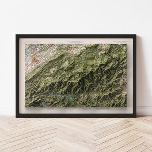 May include: A framed topographic map of a mountainous region, featuring shades of green, brown, and beige. The map is detailed with contour lines and road markings, encased in a black frame. The map is displayed on a light-colored wall.