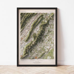 May include: A framed topographical map with a black frame. The map features a detailed relief of a mountainous region, with shades of green and brown indicating elevation. Roads and towns are marked with lines and dots.