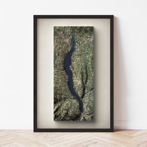 May include: A framed aerial view of a deep blue lake winding through a landscape of green and brown terrain. The artwork is encased in a black frame with a white mat, displayed against a white wall and light wood floor.