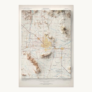 May include: A three-dimensional topographic map of Phoenix, Arizona, featuring raised relief terrain in shades of tan and brown, overlaid with a grid of red and black lines representing roads and city boundaries on a light cream background.