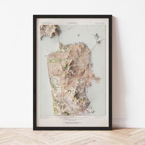 May include: Framed 3D relief map of San Francisco, California, showcasing the city's topography. The map features detailed street layouts, water features, and elevation changes. The map is framed in a black frame and is displayed on a white wall.
