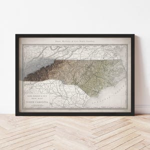 May include: Framed vintage map of North Carolina, featuring detailed cartography in muted tones of brown, green, and beige. The map is enclosed in a black frame, with text at the top and bottom indicating the map's title and publisher.
