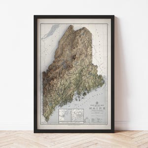 May include: Framed vintage-style map of Maine, featuring a 3D topographic representation. The map is in shades of brown and green, with the state's outline prominently displayed. The frame is black, and the map includes the text "PORT ROUTE MAP MAINE".
