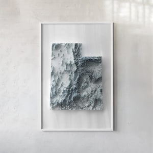 May include: A framed wall art piece featuring a three-dimensional topographic relief map of the state of Utah. The map displays detailed mountain ranges and valleys in shades of white, gray, and charcoal, encased in a simple white frame.