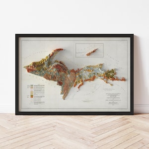 May include: Framed topographic map of the Upper Peninsula of Michigan, USA. The map features a 3D relief, showcasing the region's elevation changes with a variety of colors. The map is encased in a black frame and mounted on a white background.