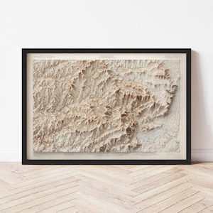 May include: A framed topographic map with a detailed relief of a mountainous region. The map features varying shades of brown and beige, with intricate contour lines and text. The black frame adds a contrasting border to the artwork.