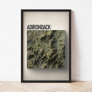 May include: A framed print featuring a topographical map of the Adirondack region. The map is rendered in shades of green, brown, and tan, with the word "ADIRONDACK" in bold black letters at the top. The print is encased in a black frame.