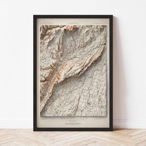 May include: Framed topographic map print with a detailed relief of a mountainous region. The map features shades of brown and beige, with a network of lines indicating roads and other features. The black frame adds a modern touch to the vintage-style artwork.