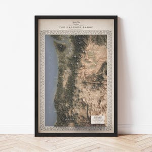 May include: Framed map print of The Cascade Range, featuring a detailed topographical view in shades of green, brown, and beige. The map includes the text "The Cascade Range" and "Atlas" with decorative borders, set against a cream-colored background.