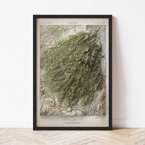 May include: A framed topographical map of a mountainous region, featuring shades of green and brown. The map is encased in a black frame, and the details include contour lines and place names. The map is displayed against a white wall and a wooden floor.