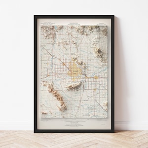 May include: Framed 3D topographic map of a city, featuring a detailed relief of the terrain. The map has a beige and tan color scheme with red and blue lines indicating roads and waterways. The map is encased in a black frame.