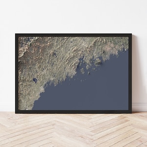 May include: Framed wall art featuring a detailed aerial view of a coastal region, likely a map. The artwork showcases a blend of natural colors, including greens, browns, and blues, within a black frame. The piece is displayed against a white wall and wooden floor.