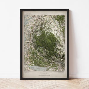 May include: A framed topographical map of a mountainous region, featuring shades of green, brown, and white. The map is encased in a black frame, adding a classic touch to the artwork. The map's details include contour lines and geographical features.