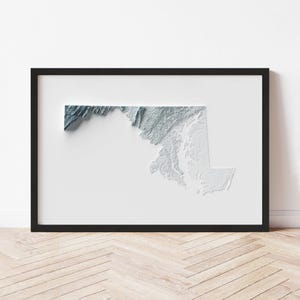 May include: A framed 3D relief map of Maryland, showcasing the state's topography in shades of blue and white. The artwork is set against a white background within a black frame, creating a striking visual contrast. The piece is displayed on a light wood floor.