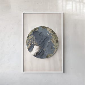 May include: A framed wall art print featuring a circular topographic map of the Arctic region. The map displays deep blue ocean depths, textured landmasses in shades of brown and green, and a prominent white ice sheet in the lower left quadrant.