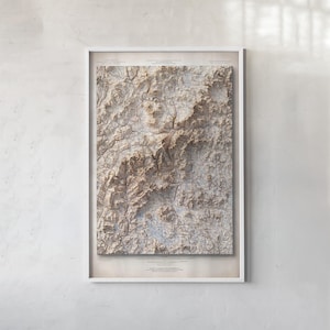 May include: A framed topographic map print featuring detailed relief shading of a mountainous region in shades of beige, brown, and light blue. The map is encased in a white frame and displays intricate terrain lines, valleys, and peaks.