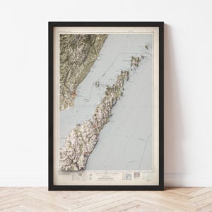 May include: A framed topographic map with a black frame. The map features a detailed relief of a coastal region, with shades of green, brown, and white. The map is set against a light blue background, with contour lines and place names.