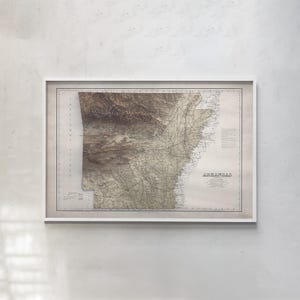 May include: A framed vintage-style map of Arkansas featuring a detailed topographical relief of the terrain in shades of brown and beige. The map includes the text ARKANSAS, various geographical labels, and a border within a white frame.