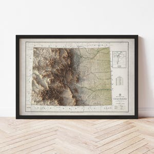 May include: Framed vintage map of Colorado in a black frame. The map features a detailed relief of the state's terrain in shades of brown and green. Includes a smaller inset map and text at the bottom. The frame is rectangular and sits on a wooden surface.