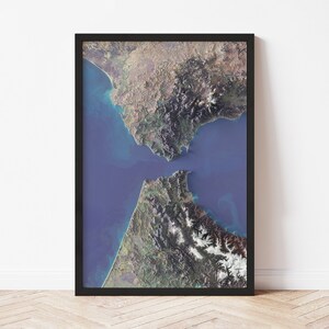 May include: A framed aerial view of a coastal landscape, showcasing land masses separated by a body of blue water. The land features varying shades of green, brown, and white, with a black frame. The image is mounted on a white wall.