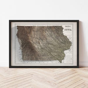 May include: Framed vintage map of Iowa, featuring detailed topographical lines and shading in shades of brown and green. The map is labeled "IOWA" in a classic font and is set against a cream-colored background, framed in black.