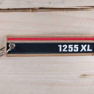 Può includere: Un portachiavi rettangolare con bordo beige, strisce rosse e nere e la scritta "1255 XL" in bianco. Ha un portachiavi in metallo argentato attaccato. Lo sfondo è una superficie di legno di colore chiaro.