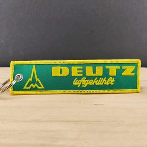 Może przedstawiać: Zielono-żółty haftowany brelok z napisem "DEUTZ" i "luftgekühlt" w kolorze żółtym. Brelok ma żółtą obwódkę i srebrny kółko na klucze. Projekt zawiera żółtą grafikę.