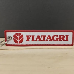 Peut inclure: Un porte-clés rectangulaire avec le texte "FIATAGRI" en rouge sur fond blanc. Il a une bordure rouge et un porte-clés argenté. Le logo FIATAGRI est également en rouge.