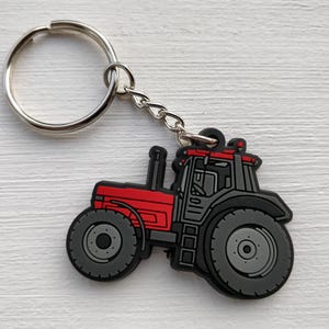 Könnte beinhalten: Ein roter und schwarzer Traktor-Schlüsselanhänger mit einem silbernen Schlüsselring und einer Kette. Der Traktor hat große graue Räder und ein detailliertes Design. Der Schlüsselanhänger ist ein Accessoire zum Thema Bauernhof.