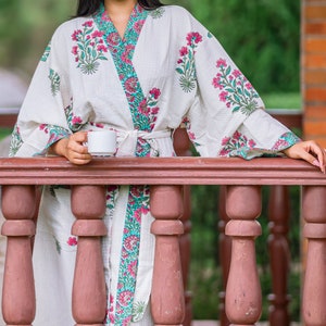 May include: A white cotton robe with pink and green floral print, tied at the waist with a teal sash. The robe is worn by a person holding a white ceramic mug.
