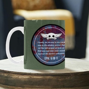 Pode incluir: Uma caneca de caf&eacute; de cer&acirc;mica verde com uma al&ccedil;a branca. A caneca apresenta um design circular com um personagem de desenho animado e texto de Ef&eacute;sios 6:10-11. O design usa cores vermelhas, brancas e azuis.