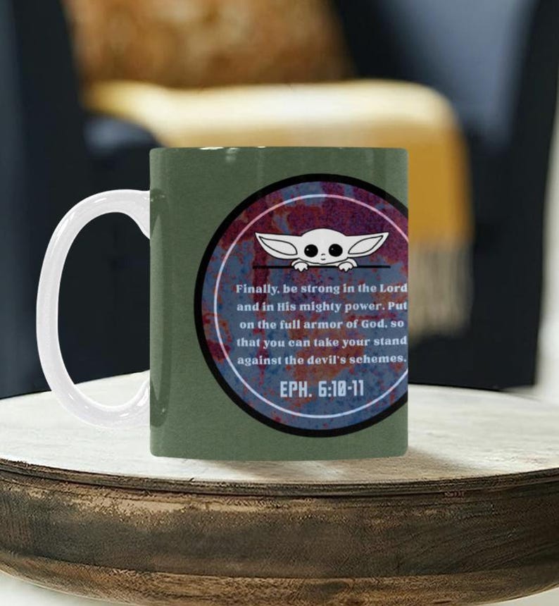 Pode incluir: Uma caneca de caf&eacute; de cer&acirc;mica verde com uma al&ccedil;a branca. A caneca apresenta um design circular com um personagem de desenho animado e texto de Ef&eacute;sios 6:10-11. O design usa cores vermelhas, brancas e azuis.