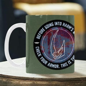 Pode incluir: Caneca de cer&acirc;mica verde com uma al&ccedil;a branca. A caneca apresenta um design circular com o texto "BEFORE GOING INTO HARM'S WAY. CHECK YOUR ARMOR. THIS IS THE WAY." e um gr&aacute;fico branco.