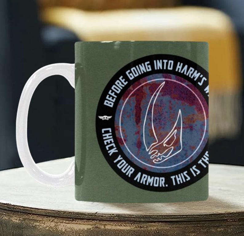 Pode incluir: Caneca de cer&acirc;mica verde com uma al&ccedil;a branca. A caneca apresenta um design circular com o texto "BEFORE GOING INTO HARM'S WAY. CHECK YOUR ARMOR. THIS IS THE WAY." e um gr&aacute;fico branco.