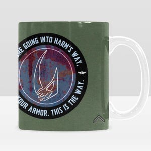 Pode incluir: Uma caneca de cer&acirc;mica verde com uma al&ccedil;a branca. A caneca apresenta um design circular com uma caveira com chifres branca e o texto "WE'RE GOING INTO HARM'S WAY. PUT ON YOUR ARMOR. THIS IS THE WAY." O design tem um aspeto envelhecido.